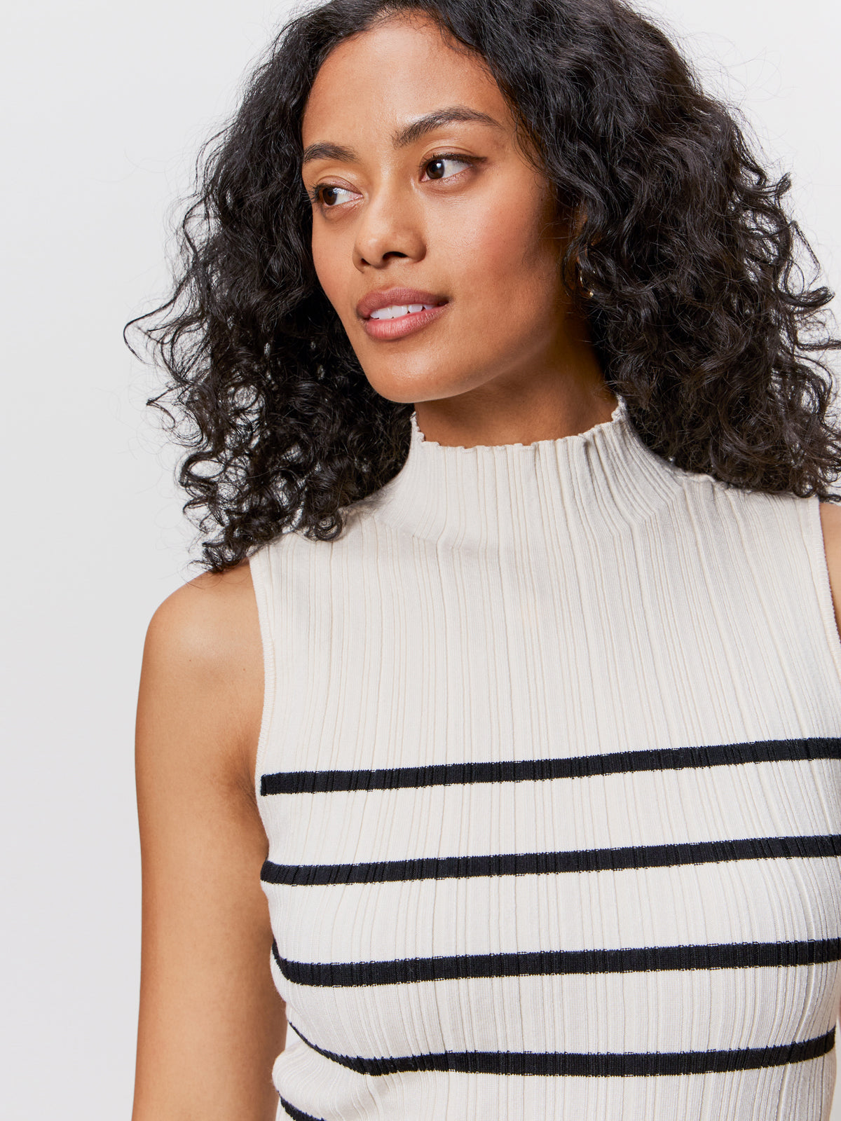 A woman with curly dark hair looks to the side, wearing the Sanctuary Clothing ribbed funnel neck top in light oat black 3. The sleeveless top features horizontal black stripes and a mock neckline against a plain white background.